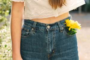 A woman, seen from waist to thigh, wears a pair of classic blue jeans with a yellow flower tucked in the pocket.