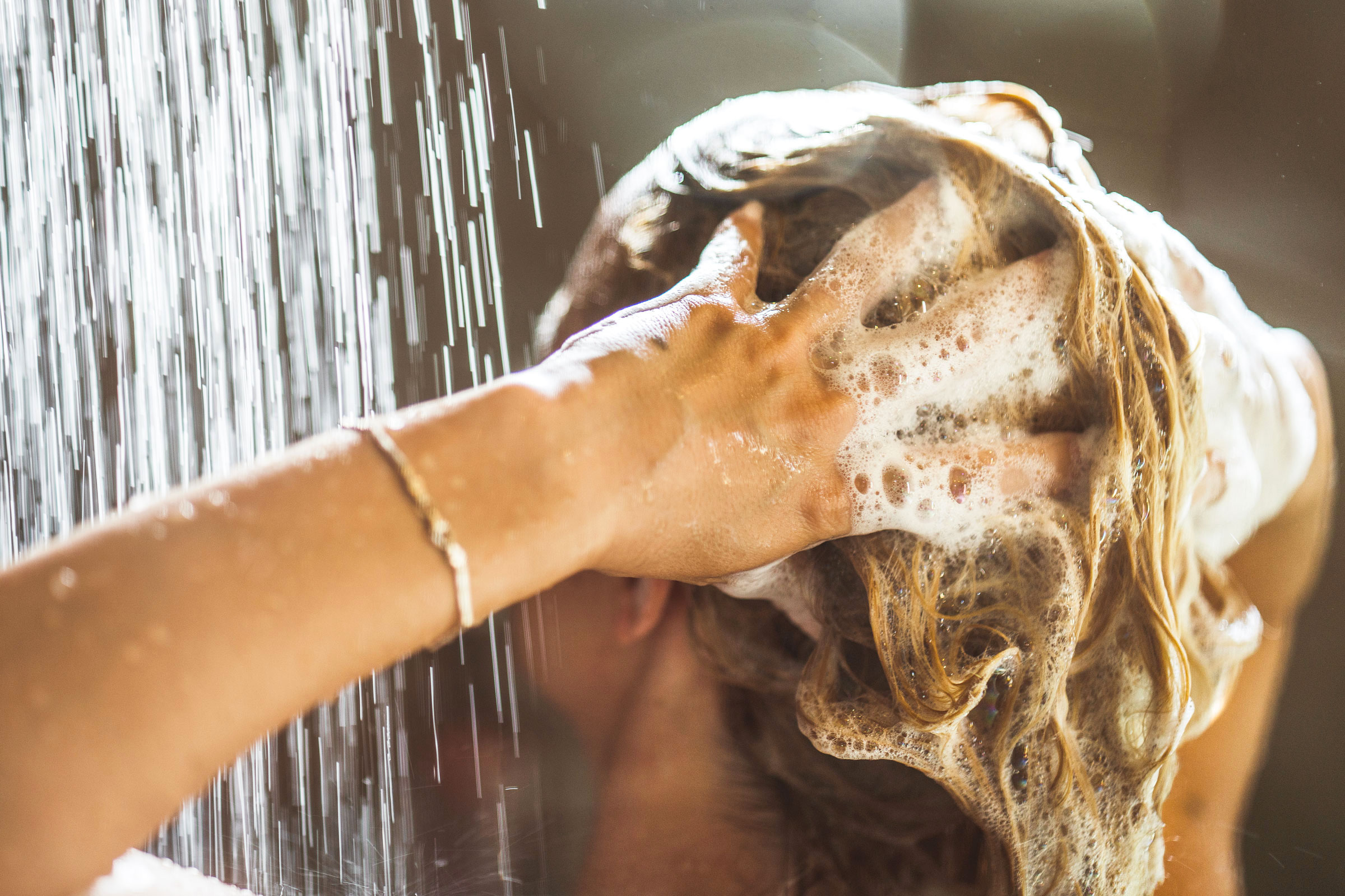 A person with long blond hair shampoos their scalp with both hands in the shower.