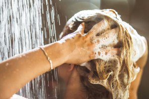 A person with long blond hair shampoos their scalp with both hands in the shower.