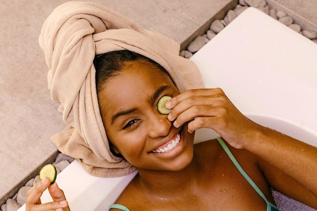 black woman laying in a bathtub with a towel on her head and a cucumber on her eye smiling 