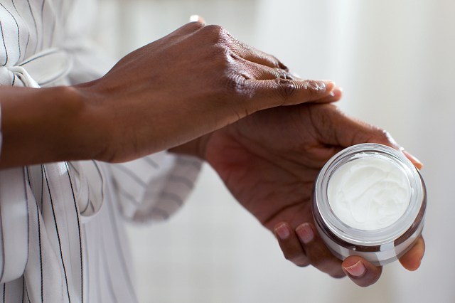 Hands of unrecognisable Black woman holding cosmetic cream and applying on her skin.