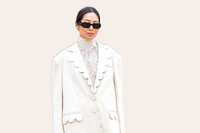 Aimee Song wears black sunglasses from Prada, white pearls earrings, a white lace print pattern high neck / long legging jumpsuit, a white linen oversized blazer jacket with wavy borders, dark brown shiny leather block heels knees boots, 
