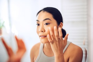 Beautiful and cheerful Asian woman standing in her bathroom, holding face cream and applying it to her cheeks and face.