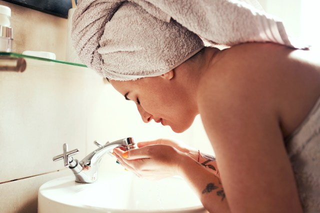 Woman washing face in sink, hair up in towel