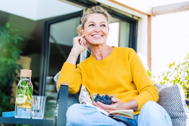 Woman in yellow sweater sitting outside porch with book and bowl of blueberries