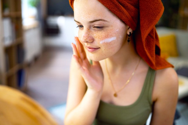 Freckled girl, with hair in towel, applying face cream in front of mirror