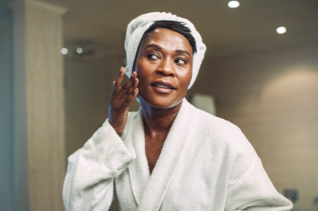 Woman in white bathrobe, towel on her head, touching her face in a bathroom