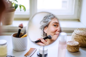 Woman applying makeup in mirror reflection with beauty products scattered around