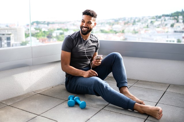 Happy, young man sitting on his roof listening to music on his phone, weights next to him