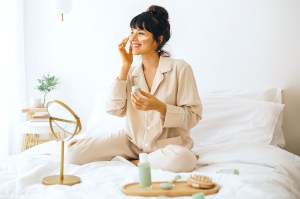 Woman sitting on bed with skin care items, putting lotion on face